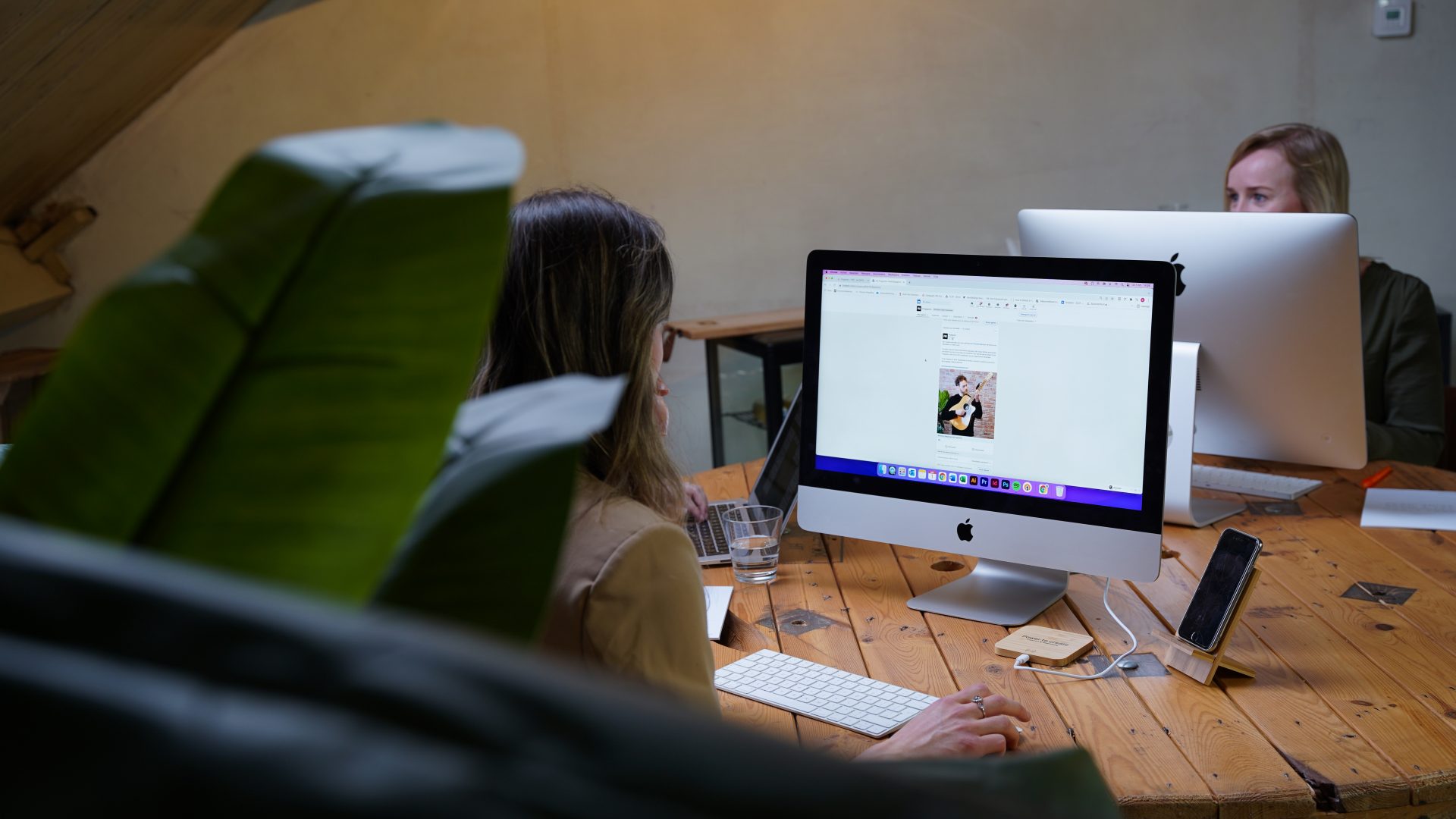 Twee vrouwen werken achter iMacs aan een houten tafel in een creatief kantoor.