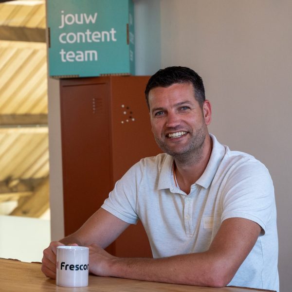 Lachende Job met Frescon-mok aan tafel in kantoorruimte.